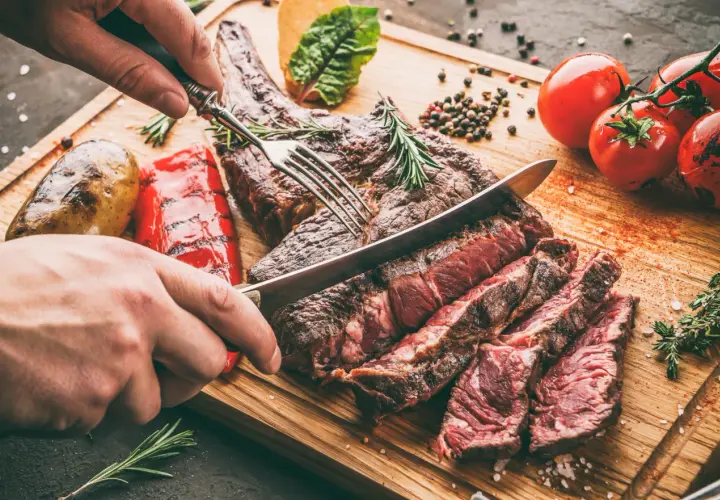 Une main tranche une côte de bœuf juteuse, résultat d'une cuisson au barbecue charbon parfaite. Accompagnée de pommes de terre et poivrons grillés.