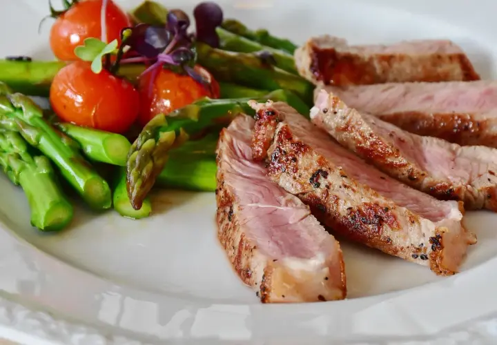 Tranches de secreto de porc grillé, accompagnées d'asperges vertes et de tomates cerises sur une assiette blanche.