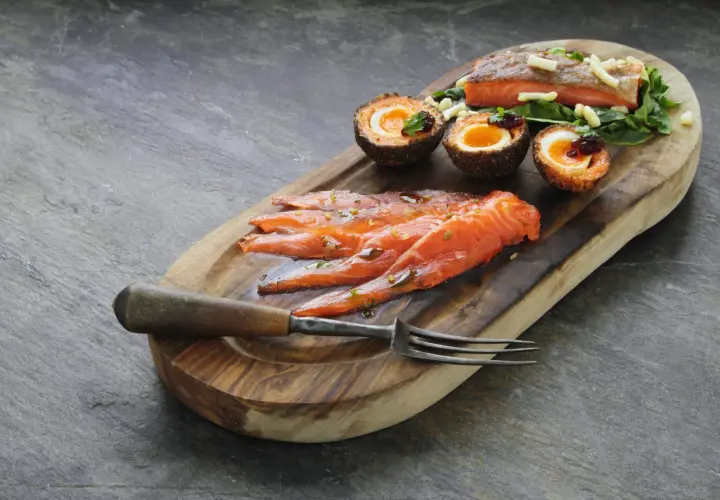 Tranché de truite fumée, œufs panés et garniture sur planche en bois. La cuisson truite fumée est mise en valeur.