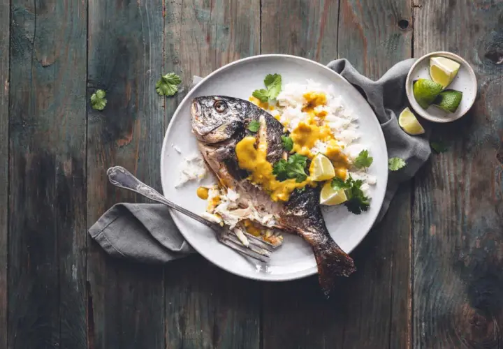 Dorade grise entière, parfaitement cuite, servie avec du riz blanc, une sauce jaune onctueuse, des quartiers de citron vert et de la coriandre fraîche. Un délice visuel pour la cuisson dorade grise.