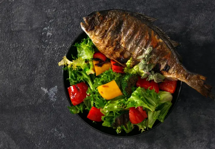 Dorade au barbecue, entière et grillée, servie sur un lit de salade verte, poivrons rouges et jaunes, et brocolis.