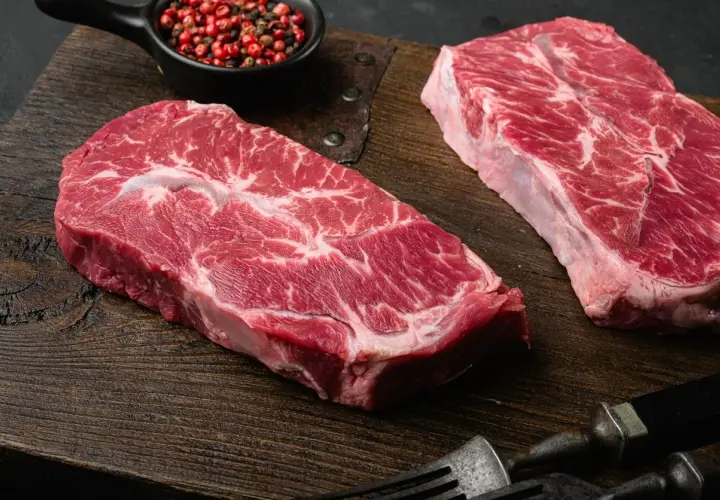 Deux beaux steaks de l angus beef, marbrés de gras, prêts à être cuisinés sur une planche en bois. Poivre rose en accompagnement.