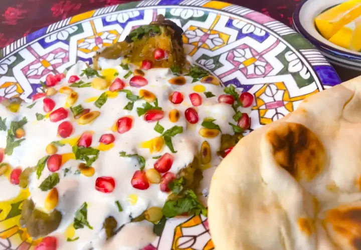 Un plat de mutabal crémeux, garni de grenades et de pignons, servi avec du pain pita sur une assiette décorée.