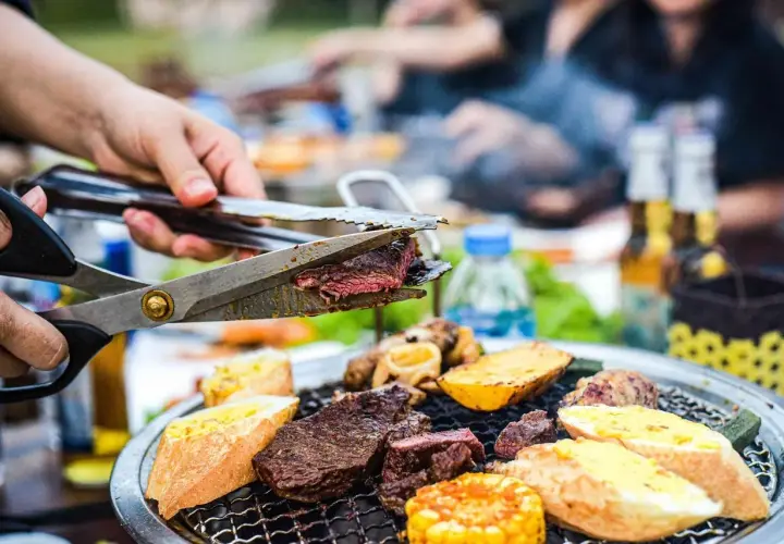 Une main coupe un steak juteux sur un gril lors d'une fête barbecue. Des pains grillés, des pommes de terre et du maïs complètent le festin.