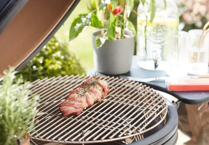 Un rôti de viande enroulé de lard, prêt à griller. Une légère **fume bleu** s'échappe, promettant un repas savoureux en plein air.