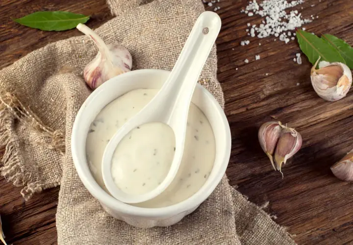 Sauce à l'ail crémeuse dans un bol blanc, avec une cuillère, des gousses d'ail et des feuilles de laurier sur une table en bois rustique.