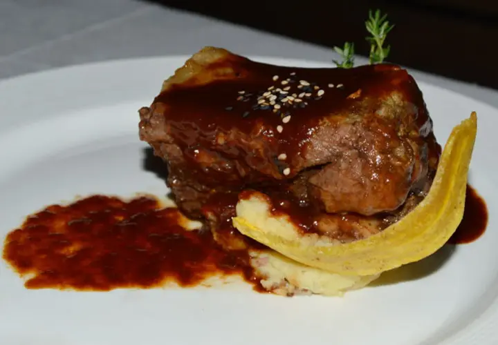 Un plat de ribs de boeuf barbecue, nappé d'une sauce foncée, servi avec une purée et une tranche de banane plantain frite.