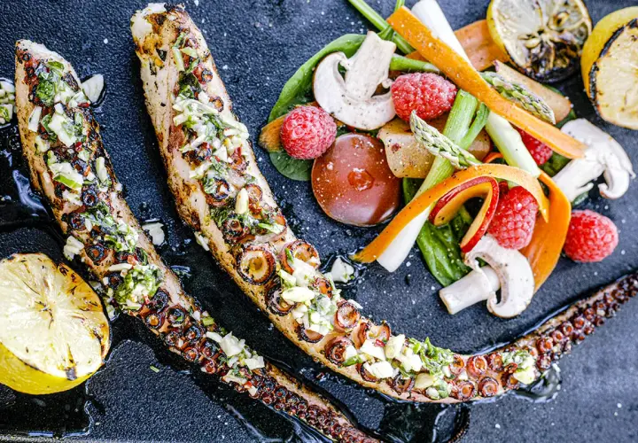 Tentacules de poulpe au barbecue, grillés et garnis d'herbes fraîches, servis avec des légumes croquants et des quartiers de citron grillé.