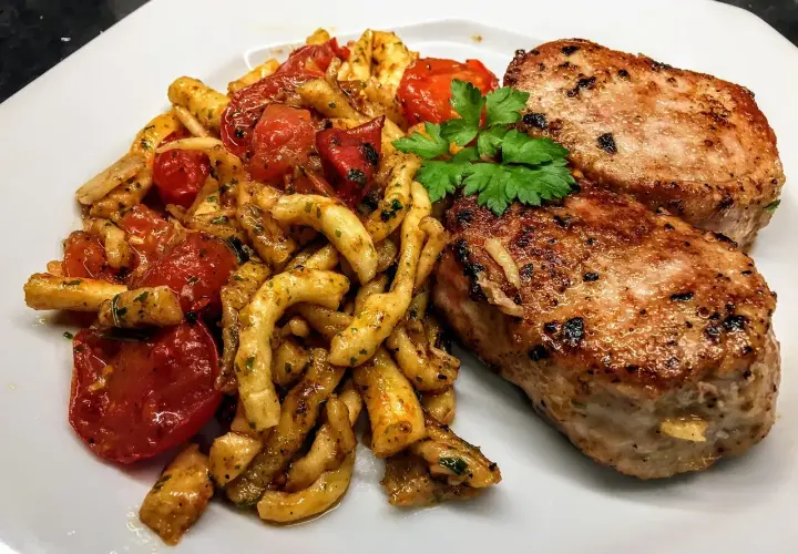 Deux côtelettes de veau grillées, servies avec des pâtes torsadées et des tomates cerises poêlées, garnies de persil frais. Un plat savoureux et coloré.