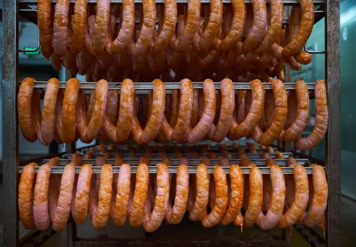 Des saucisses roses et orangées pendent en rangées sur des étagères métalliques, prêtes pour le processus de fumer des saucisses à froid.