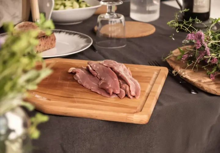 Aiguillettes de canard crues sur une planche à découper en bois, prêtes à être cuisinées. Des herbes fraîches et du pain complètent la scène.