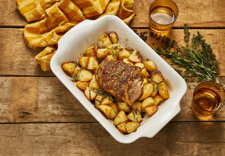 Selle d'agneau rôtie avec pommes de terre dorées et herbes aromatiques dans un plat blanc, prête à être dégustée.