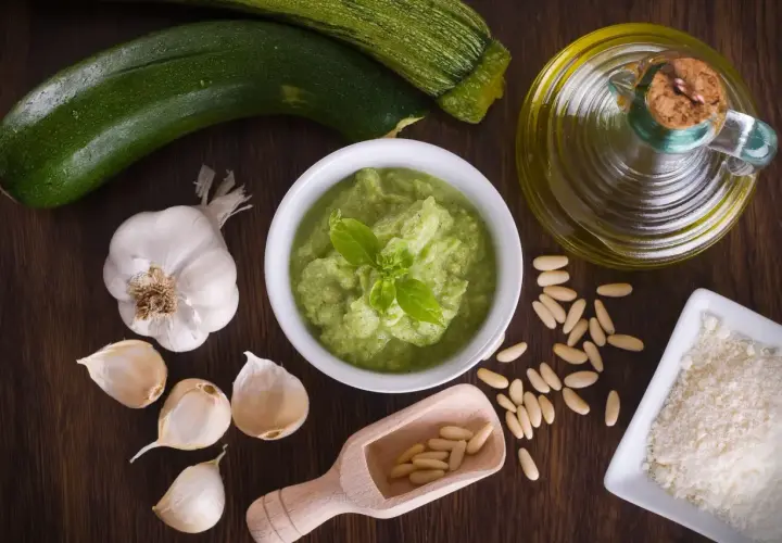 Un bol de pesto de courgettes frais, garni de basilic, entouré d'ingrédients : courgettes, ail, pignons de pin, parmesan râpé et huile d'olive.