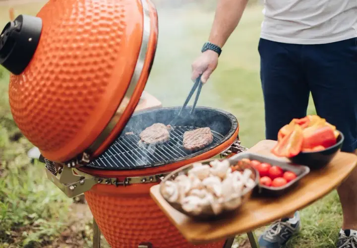 Un homme cuisine des steaks sur un kamado, c'est quoi ce barbecue en céramique orange. Des légumes sont prêts à être grillés.