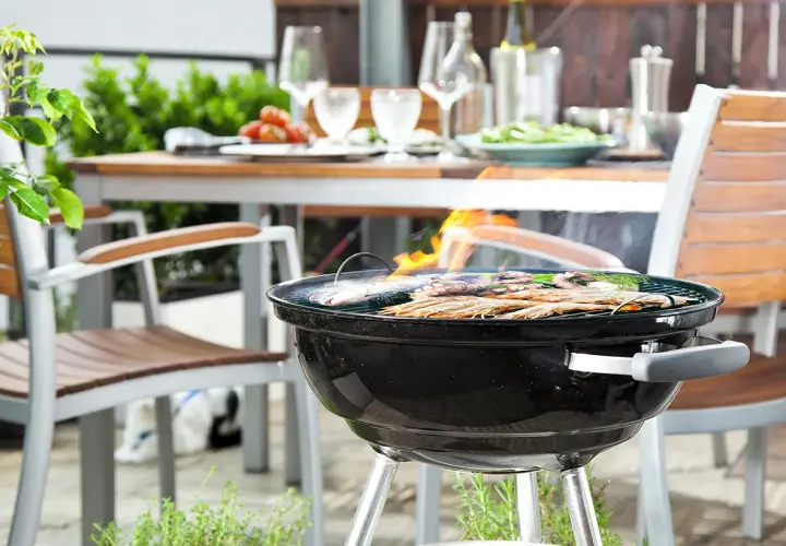 Un barbecue au charbon est allumé, avec des crevettes et du poisson grillant sur la grille. Une table est dressée à l'arrière, prête pour un repas.