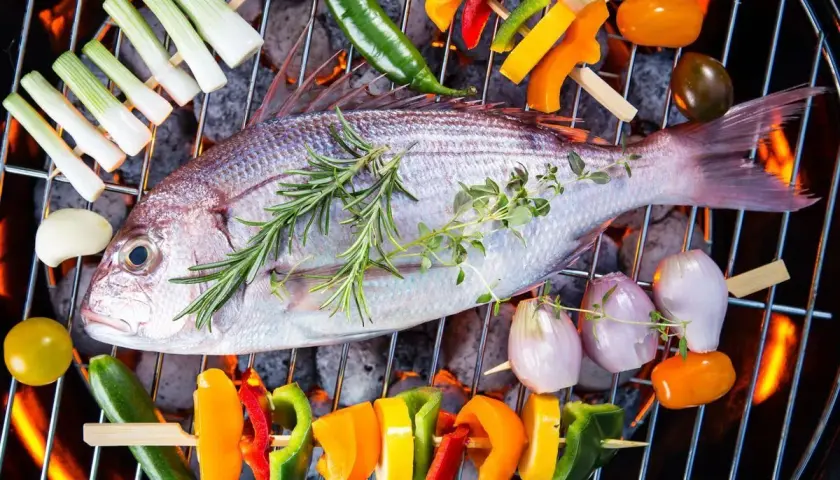 Poisson entier sur un barbecue électrique, entouré de brochettes de légumes colorés et d'herbes aromatiques.