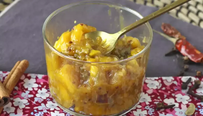 Un verre rempli de chutney de mangue, avec une cuillère dorée. Des épices comme la cannelle, le piment et le clou de girofle entourent le plat.