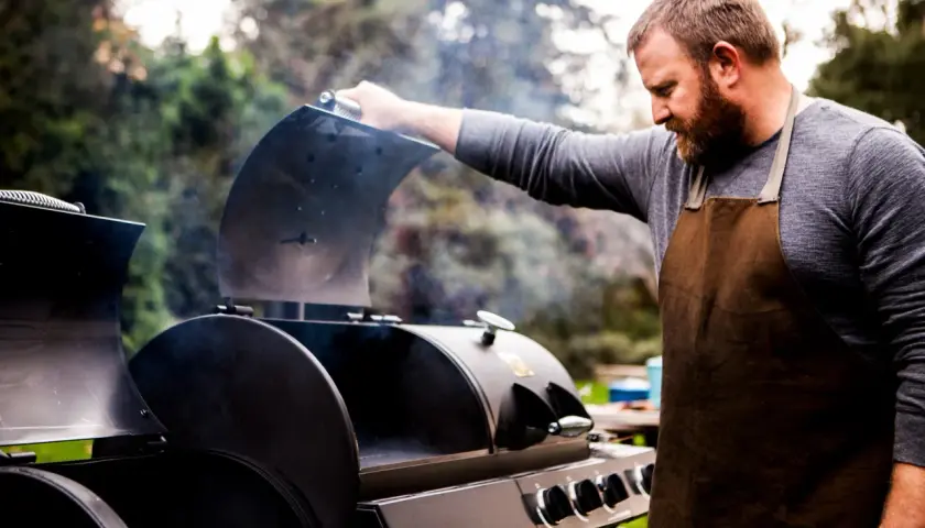 Un homme barbu en tablier ouvre un fumoir d'où s'échappe de la fumée. Il est prêt à apprendre comment allumer un fumoir pour un repas savoureux.
