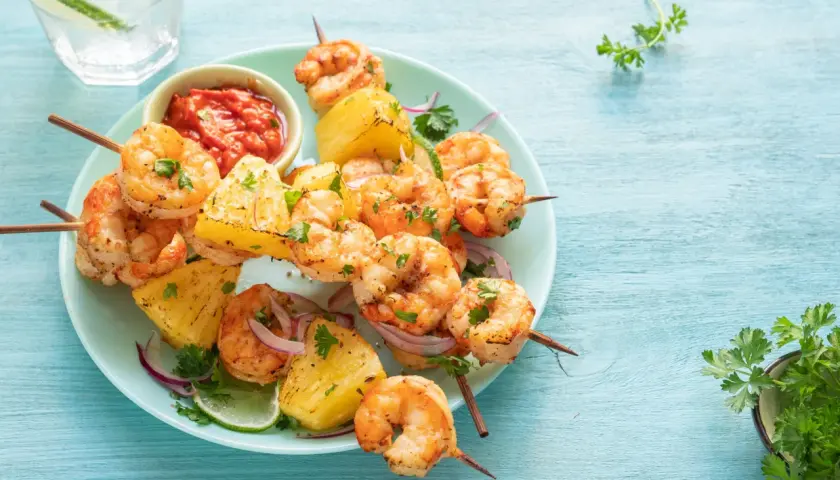 Brochettes de scampis grillés avec des morceaux d'ananas, de la ciboulette et une sauce rouge épicée sur une assiette bleue.