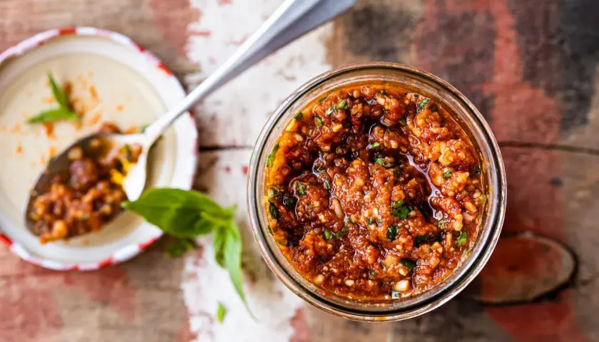 Un pot de pesto rosso, aux tomates séchées et basilic, prêt à être dégusté. Une cuillère en argent en est remplie.