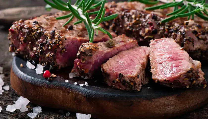 Un succulent filet mignon basse température, coupé en tranches, garni de romarin et de poivre concassé, sur une planche en bois rustique.