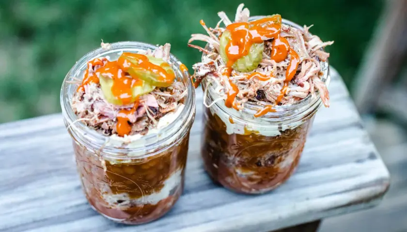 Deux pots en verre remplis de pulled pork, haricots et sauce barbecue, parfaits pour un brunch barbecue en plein air.