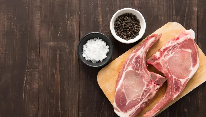 Deux belles côtes de porc crues sur une planche à découper en bois, accompagnées de sel et de grains de poivre noir.