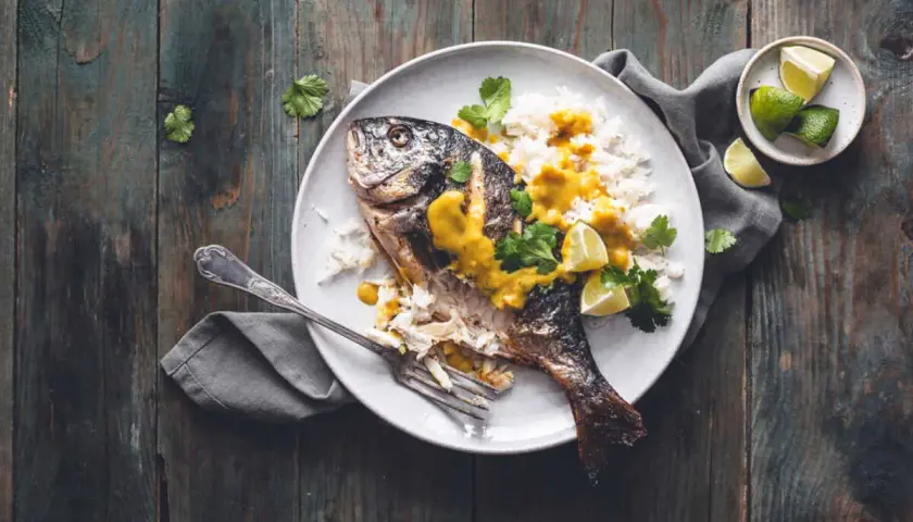 Dorade grise entière, parfaitement cuite, servie avec du riz blanc, une sauce jaune onctueuse, des quartiers de citron vert et de la coriandre fraîche. Un délice visuel pour la cuisson dorade grise.