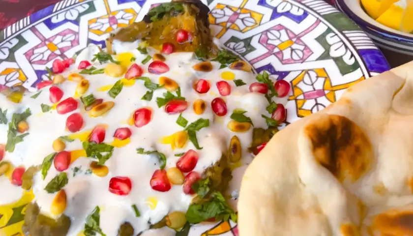 Un plat de mutabal crémeux, garni de grenades et de pignons, servi avec du pain pita sur une assiette décorée.