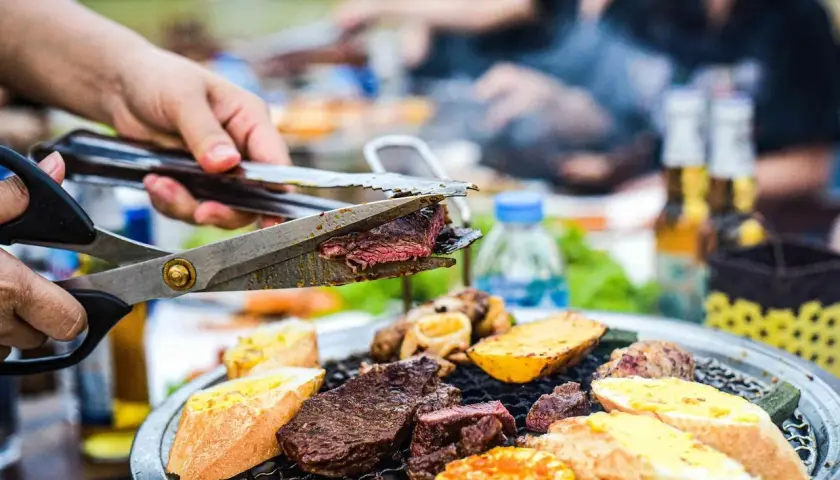 Une main coupe un steak juteux sur un gril lors d'une fête barbecue. Des pains grillés, des pommes de terre et du maïs complètent le festin.