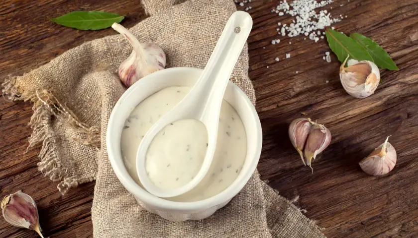 Sauce à l'ail crémeuse dans un bol blanc, avec une cuillère, des gousses d'ail et des feuilles de laurier sur une table en bois rustique.