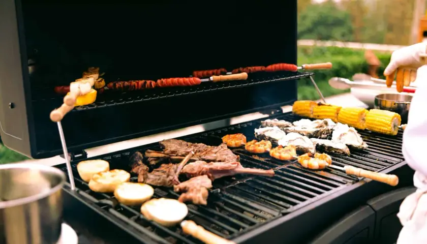 Viande, crevettes, huîtres et maïs grillés sur un barbecue gaz ou charbon. Des saucisses enroulées cuisent sur la grille supérieure.