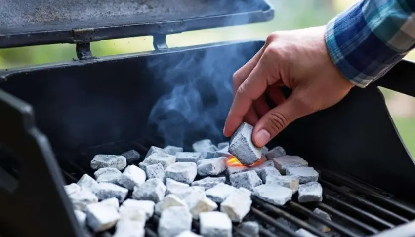 Une main allume des briquettes de charbon dans un barbecue, de la fumée s'échappe. On se demande combien de temps pour allumer un barbecue.