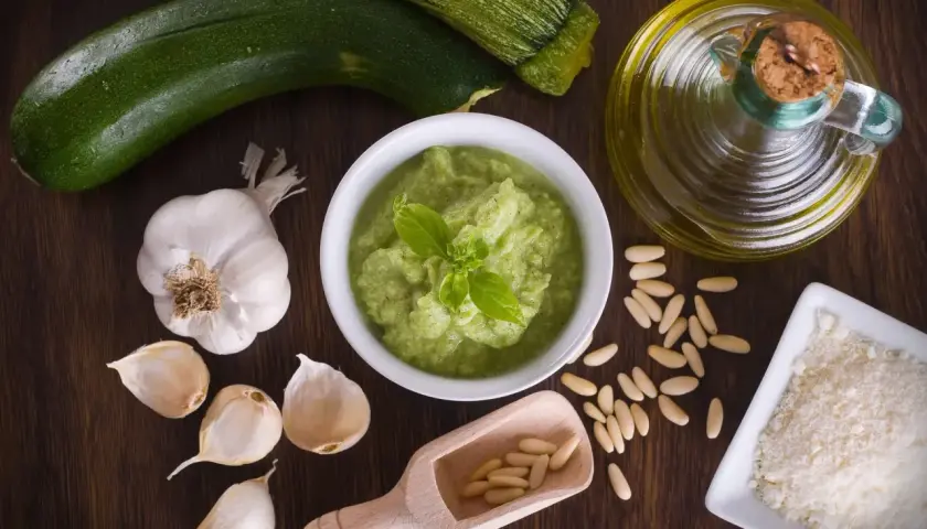 Un bol de pesto de courgettes frais, garni de basilic, entouré d'ingrédients : courgettes, ail, pignons de pin, parmesan râpé et huile d'olive.
