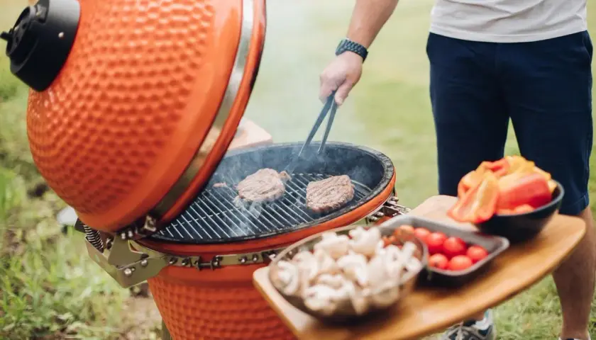 Un homme cuisine des steaks sur un kamado, c'est quoi ce barbecue en céramique orange. Des légumes sont prêts à être grillés.