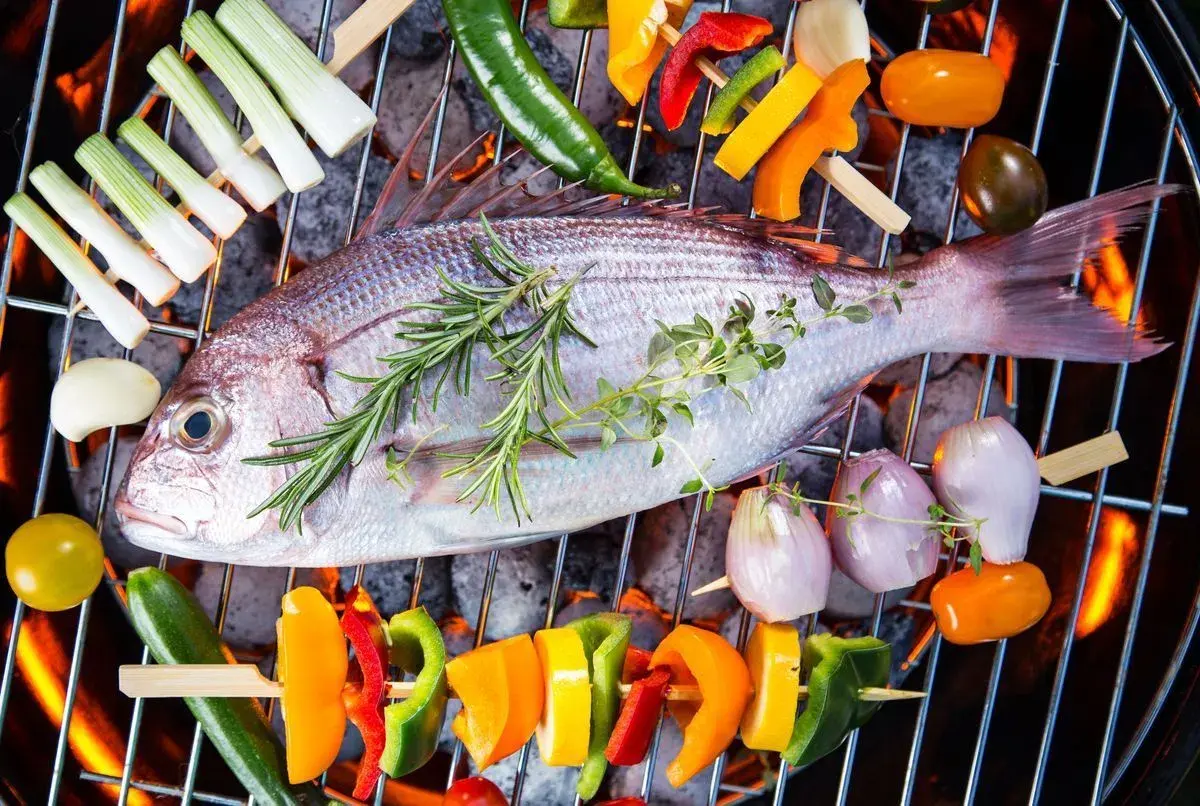 Poisson entier sur un barbecue électrique, entouré de brochettes de légumes colorés et d'herbes aromatiques.