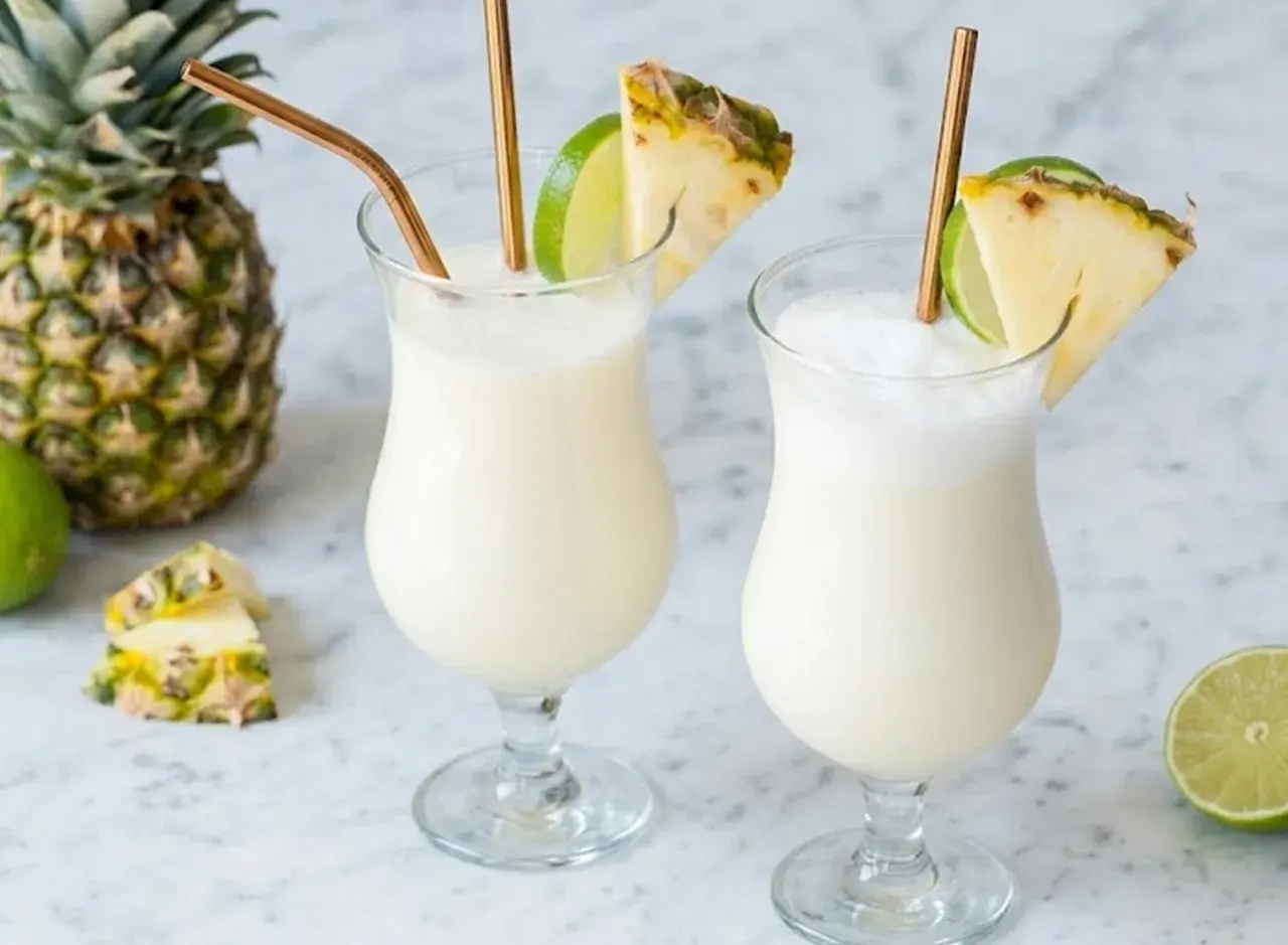 Deux verres de pina colada, décorés d'ananas et de citron vert, avec des pailles dorées. Un ananas entier et des citrons verts sont disposés autour.