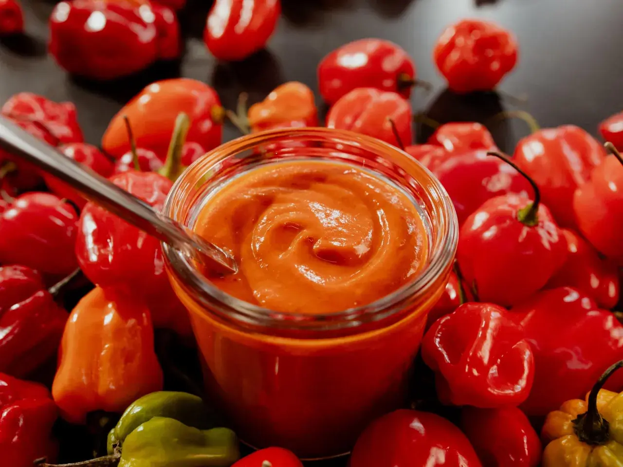 Un pot de sauce piquante maison, entouré de piments rouges et verts. Une cuillère est plongée dans la sauce, prête à être dégustée. Une recette de sauce piquante à essayer !
