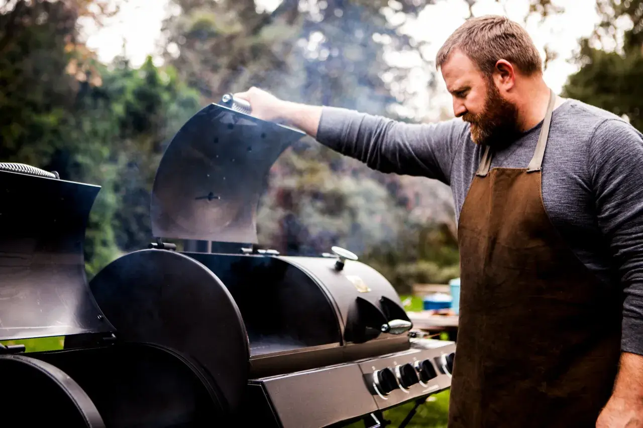 Un homme barbu en tablier ouvre un fumoir d'où s'échappe de la fumée. Il est prêt à apprendre comment allumer un fumoir pour un repas savoureux.