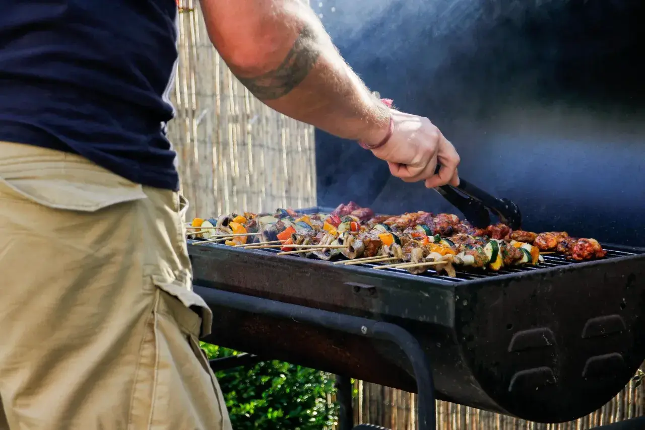 Un homme utilise des pinces pour retourner des brochettes colorées sur un barbecue fumant. C'est le moment idéal pour degraisser le barbecue après ce festin !
