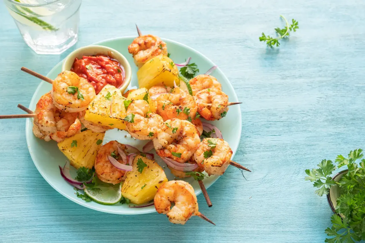Brochettes de scampis grillés avec des morceaux d'ananas, de la ciboulette et une sauce rouge épicée sur une assiette bleue.