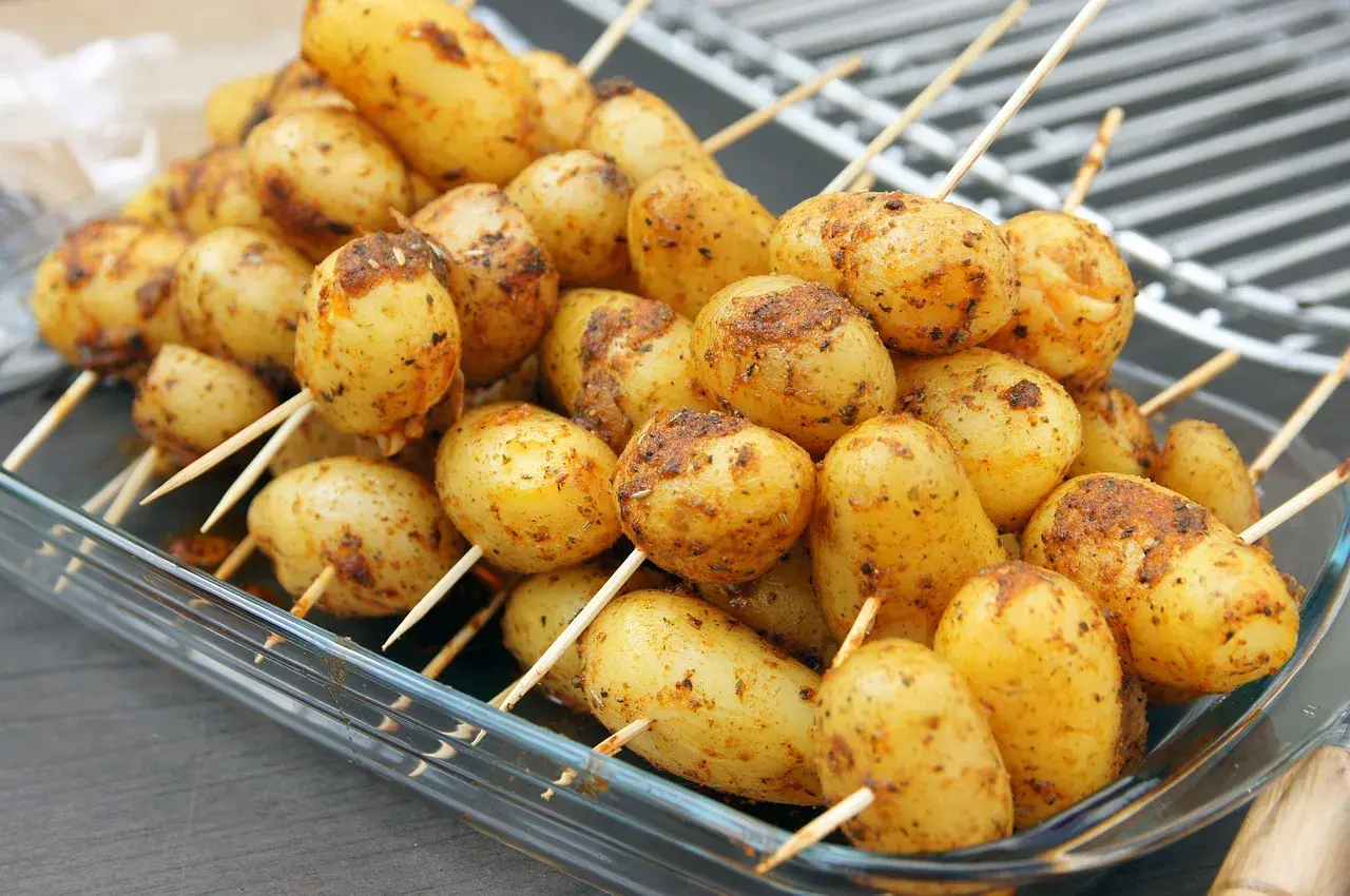 Des brochettes de pommes de terre assaisonnées, prêtes pour le barbecue, dans un plat en verre.