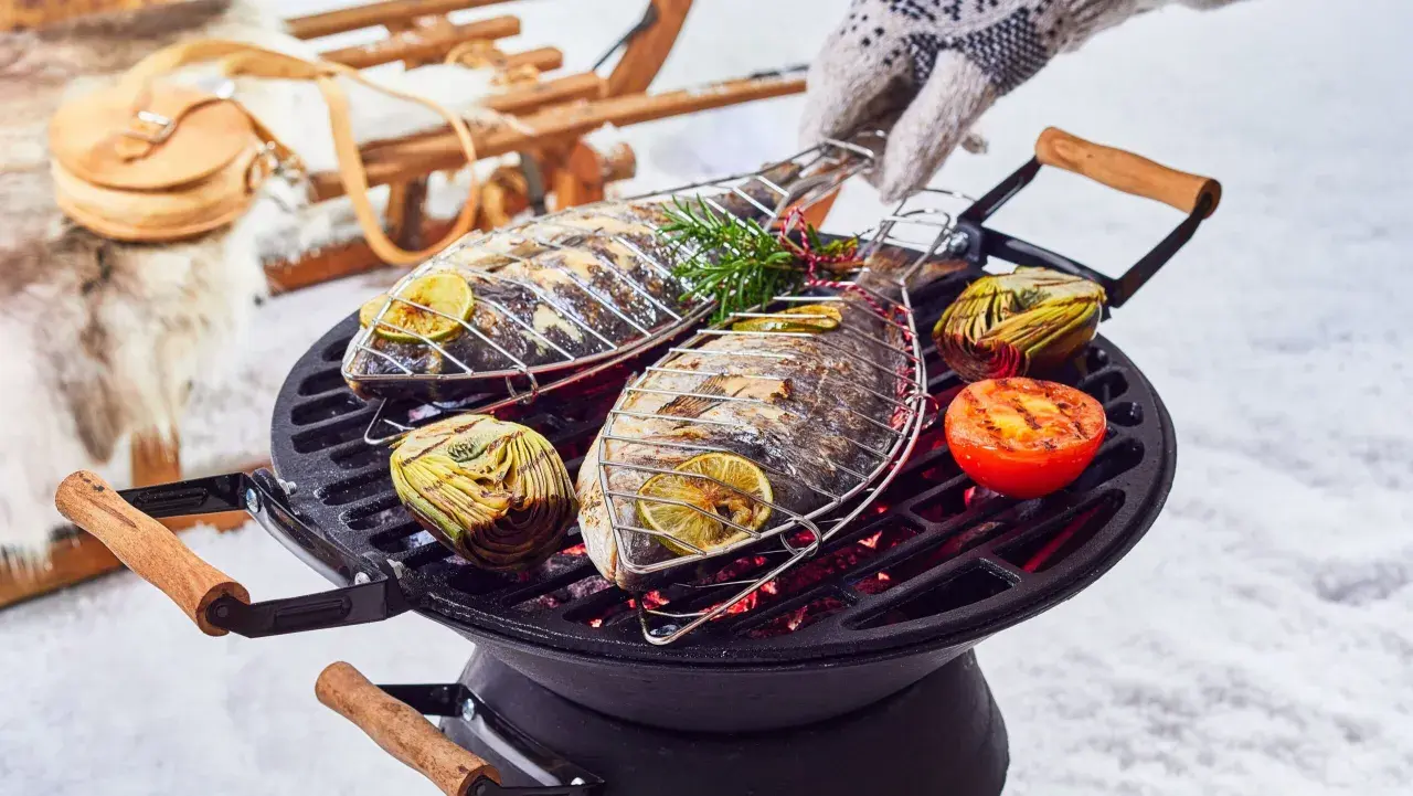 Deux poissons entiers grillent sur un barbecue, accompagnés d'artichauts et de tomates. Un gant tricoté ajuste la grille. Un vrai barbecue hivernal !