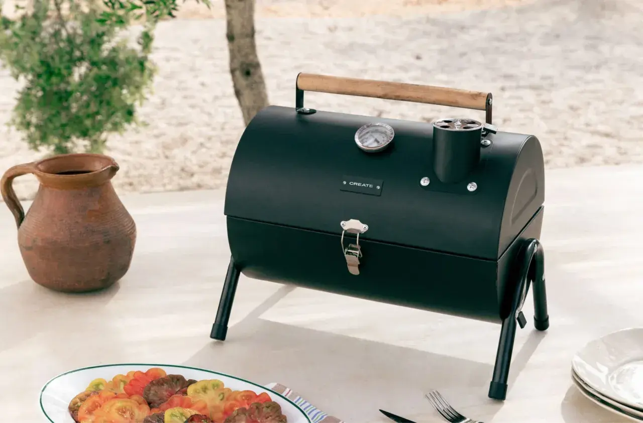 Barbecue noir avec couvercle, thermomètre et poignée en bois, prêt pour le temps de cuisson. Une carafe en terre cuite et une assiette de tomates colorées sont à proximité.