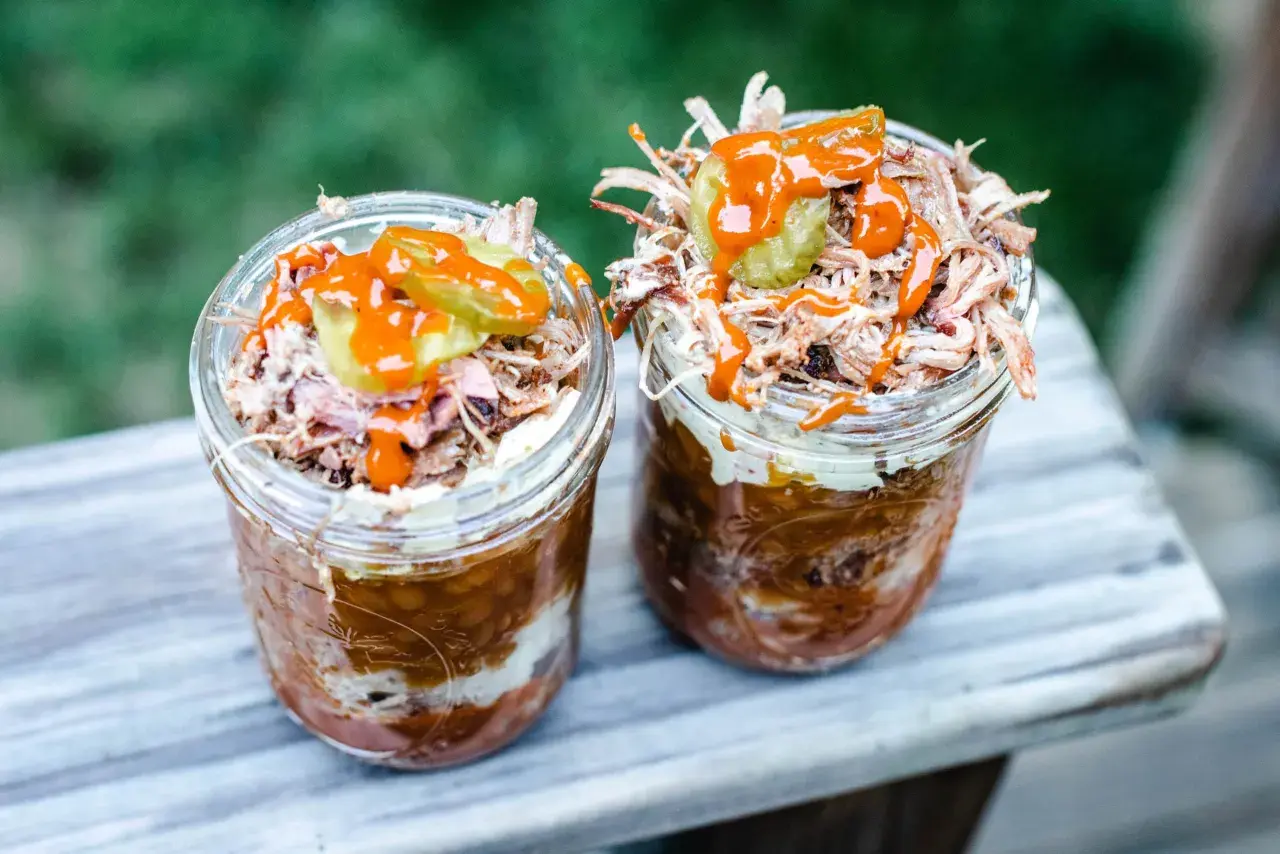 Deux pots en verre remplis de pulled pork, haricots et sauce barbecue, parfaits pour un brunch barbecue en plein air.