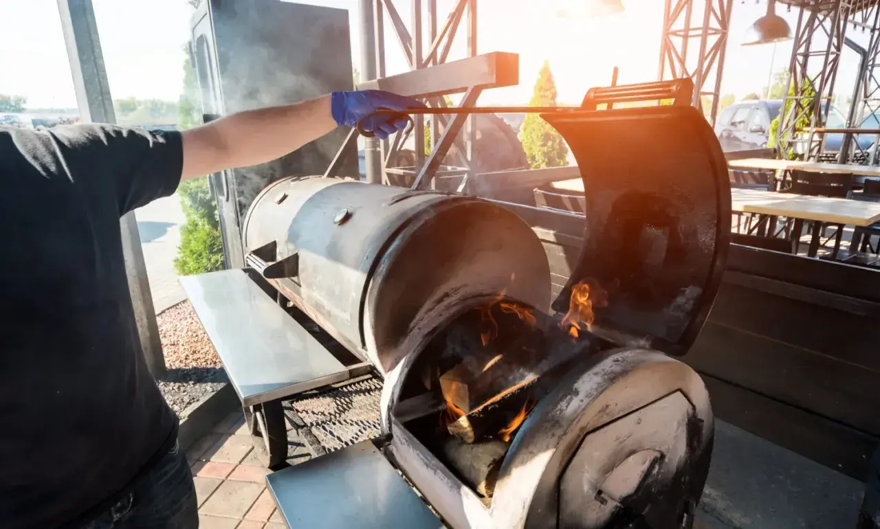 Un homme ajuste le bois pour fumoir dans un grand barbecue. Des flammes dansent dans le foyer, promettant une cuisson savoureuse.