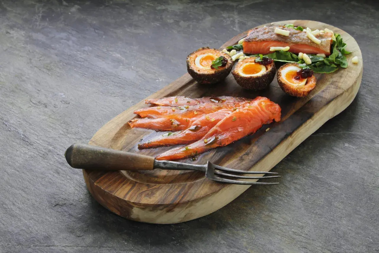 Tranché de truite fumée, œufs panés et garniture sur planche en bois. La cuisson truite fumée est mise en valeur.