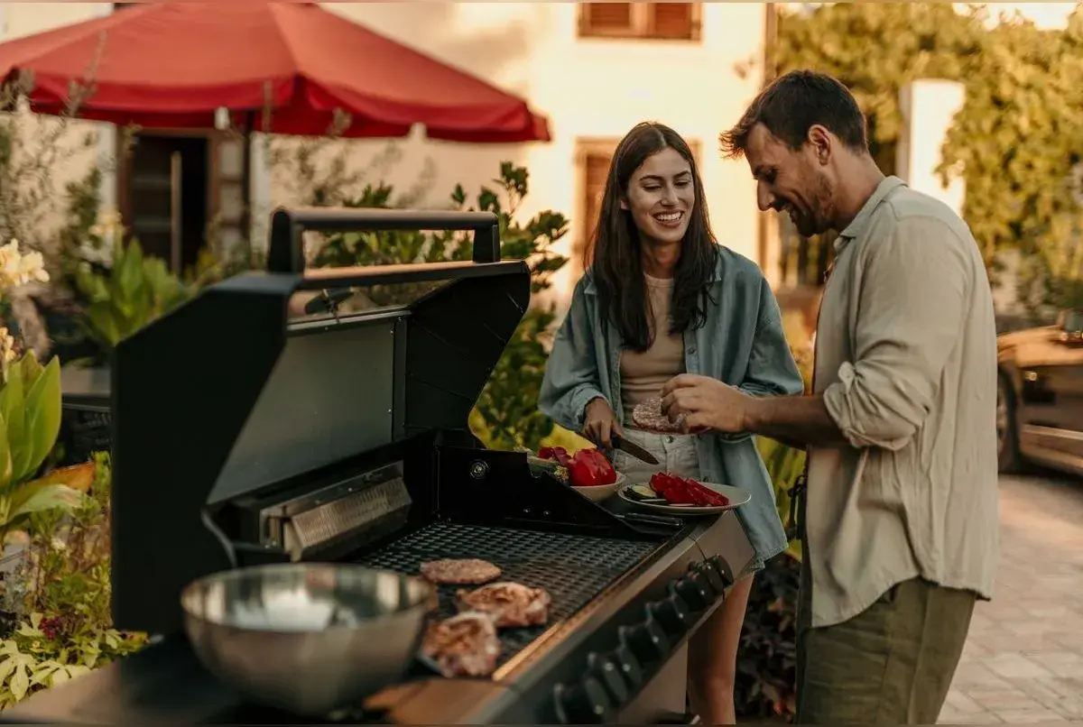 Un couple prépare une soiree barbecue party. Des steaks cuisent sur le grill pendant qu'ils coupent des légumes.