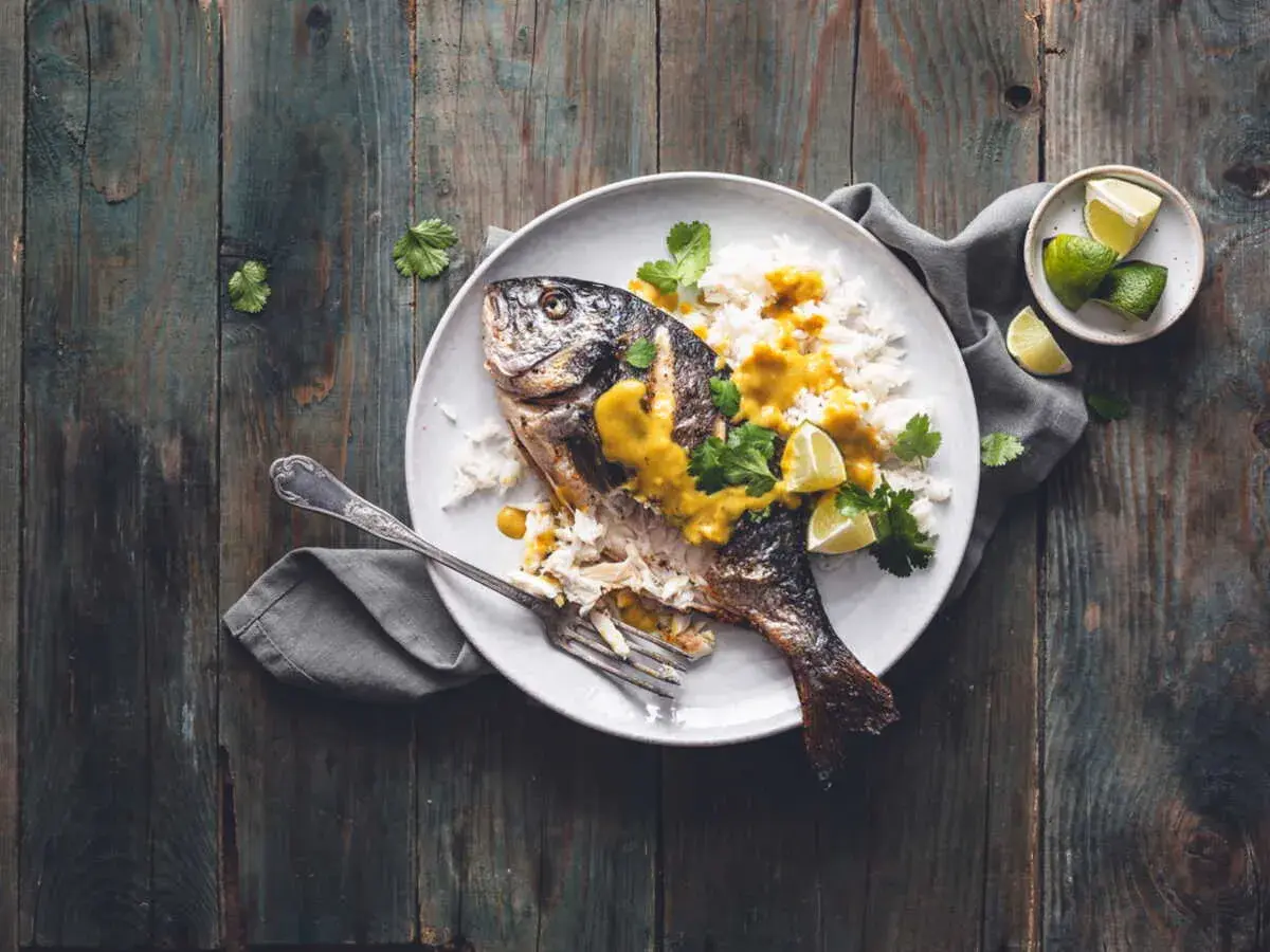 Dorade grise entière, parfaitement cuite, servie avec du riz blanc, une sauce jaune onctueuse, des quartiers de citron vert et de la coriandre fraîche. Un délice visuel pour la cuisson dorade grise.