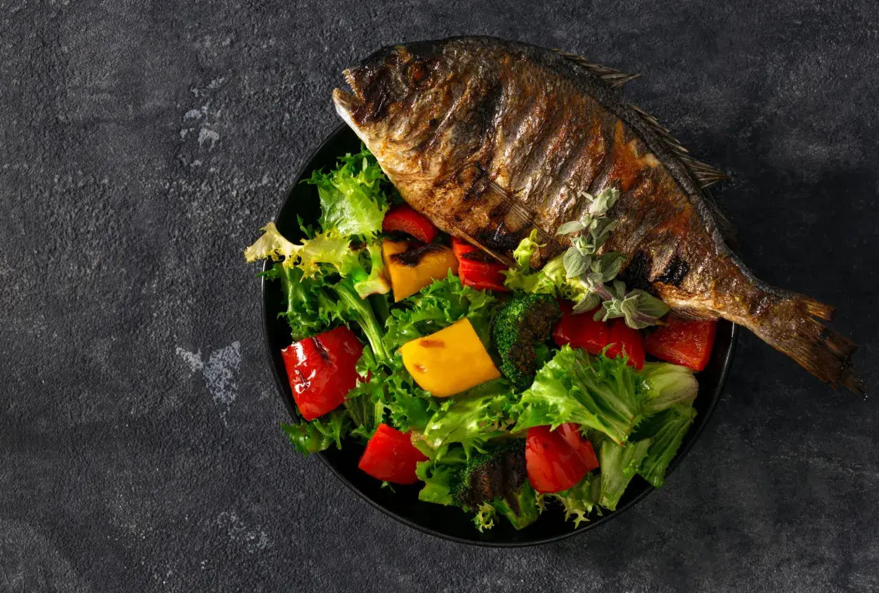 Dorade au barbecue, entière et grillée, servie sur un lit de salade verte, poivrons rouges et jaunes, et brocolis.